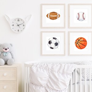 May include: Four framed watercolor paintings of different sports balls: a football, a baseball, a soccer ball, and a basketball. The paintings are hung on a white wall in a nursery setting.