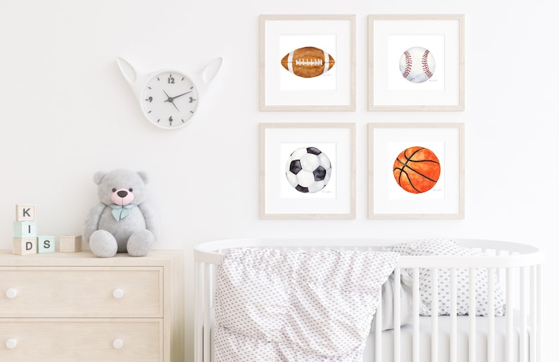 May include: Four framed watercolor paintings of different sports balls: a football, a baseball, a soccer ball, and a basketball. The paintings are hung on a white wall in a nursery setting.