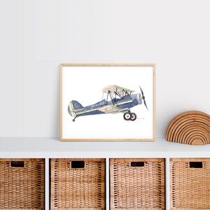 Puede incluir: Una pintura de acuarela de un biplano azul y verde sobre un fondo blanco. El avi&oacute;n est&aacute; en vuelo y tiene una h&eacute;lice en la parte delantera. La pintura est&aacute; enmarcada en un marco de madera claro.