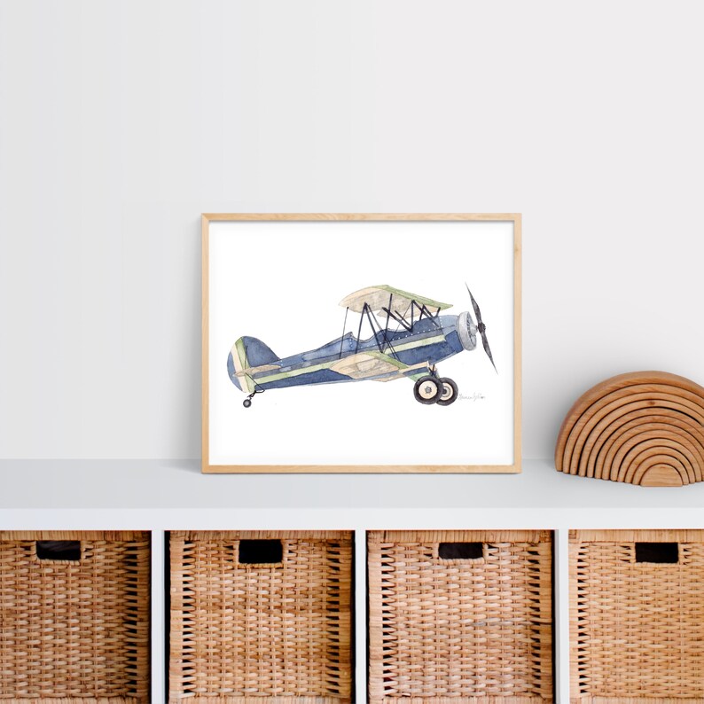 Puede incluir: Una pintura de acuarela de un biplano azul y verde sobre un fondo blanco. El avi&oacute;n est&aacute; en vuelo y tiene una h&eacute;lice en la parte delantera. La pintura est&aacute; enmarcada en un marco de madera claro.