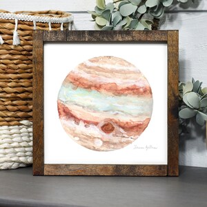 Peut inclure: Peinture &agrave; l'aquarelle de Jupiter dans un cadre en bois fonc&eacute;. La plan&egrave;te est repr&eacute;sent&eacute;e avec des bandes de couleurs orange, marron et bleu clair. L'&oelig;uvre est sign&eacute;e dans le coin inf&eacute;rieur droit. La peinture est expos&eacute;e sur une &eacute;tag&egrave;re avec un panier tress&eacute; et de la verdure d&eacute;corative.