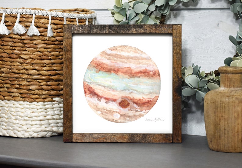 Peut inclure: Peinture &agrave; l'aquarelle de Jupiter dans un cadre en bois fonc&eacute;. La plan&egrave;te est repr&eacute;sent&eacute;e avec des bandes de couleurs orange, marron et bleu clair. L'&oelig;uvre est sign&eacute;e dans le coin inf&eacute;rieur droit. La peinture est expos&eacute;e sur une &eacute;tag&egrave;re avec un panier tress&eacute; et de la verdure d&eacute;corative.