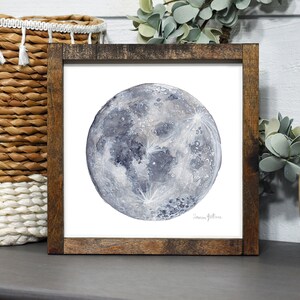 K&ouml;nnte beinhalten: Ein gerahmtes Aquarellbild eines Vollmonds vor wei&szlig;em Hintergrund. Der Mond ist in Graut&ouml;nen und Blau dargestellt, mit einer detaillierten Darstellung seiner Oberfl&auml;che.