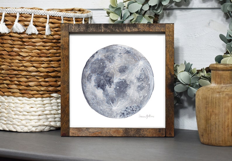 K&ouml;nnte beinhalten: Ein gerahmtes Aquarellbild eines Vollmonds vor wei&szlig;em Hintergrund. Der Mond ist in Graut&ouml;nen und Blau dargestellt, mit einer detaillierten Darstellung seiner Oberfl&auml;che.