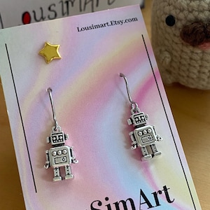 May include: Silver robot earrings with a dangling design. The earrings are displayed on a card with the text "LouSimArt.Etsy.com" and a gradient background. A gold star stud is also visible.