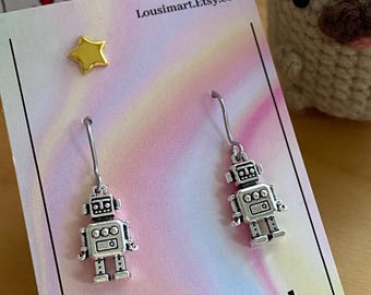May include: A pair of silver robot drop earrings. The earrings are presented on a card with the text "LouSimArt.Etsy.com" and a pastel gradient background. A gold star stud is also visible.