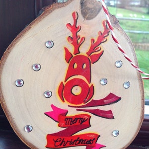 May include: A wooden Christmas ornament with a red reindeer painted on it. The reindeer has a red nose and antlers. The ornament also has a red and pink ribbon with the words "merry Christmas" written on it.