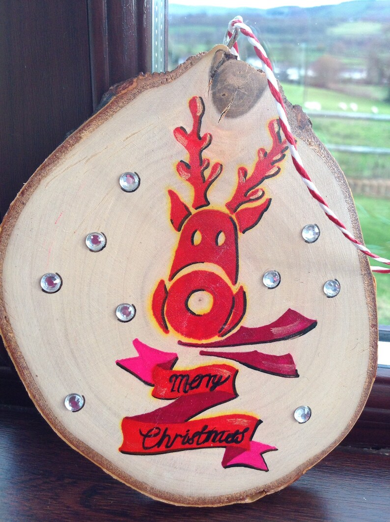 May include: A wooden Christmas ornament with a red reindeer painted on it. The reindeer has a red nose and antlers. The ornament also has a red and pink ribbon with the words "merry Christmas" written on it.