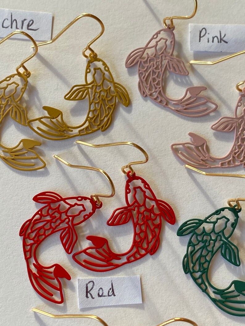 K&ouml;nnte beinhalten: Ein Set mit f&uuml;nf Paaren goldfarbener fischf&ouml;rmiger Ohrringe. Die Ohrringe sind in Rot, Rosa, Gr&uuml;n und Gold erh&auml;ltlich. Die Ohrringe sind mit "Rot", "Rosa" und "Gold" beschriftet.
