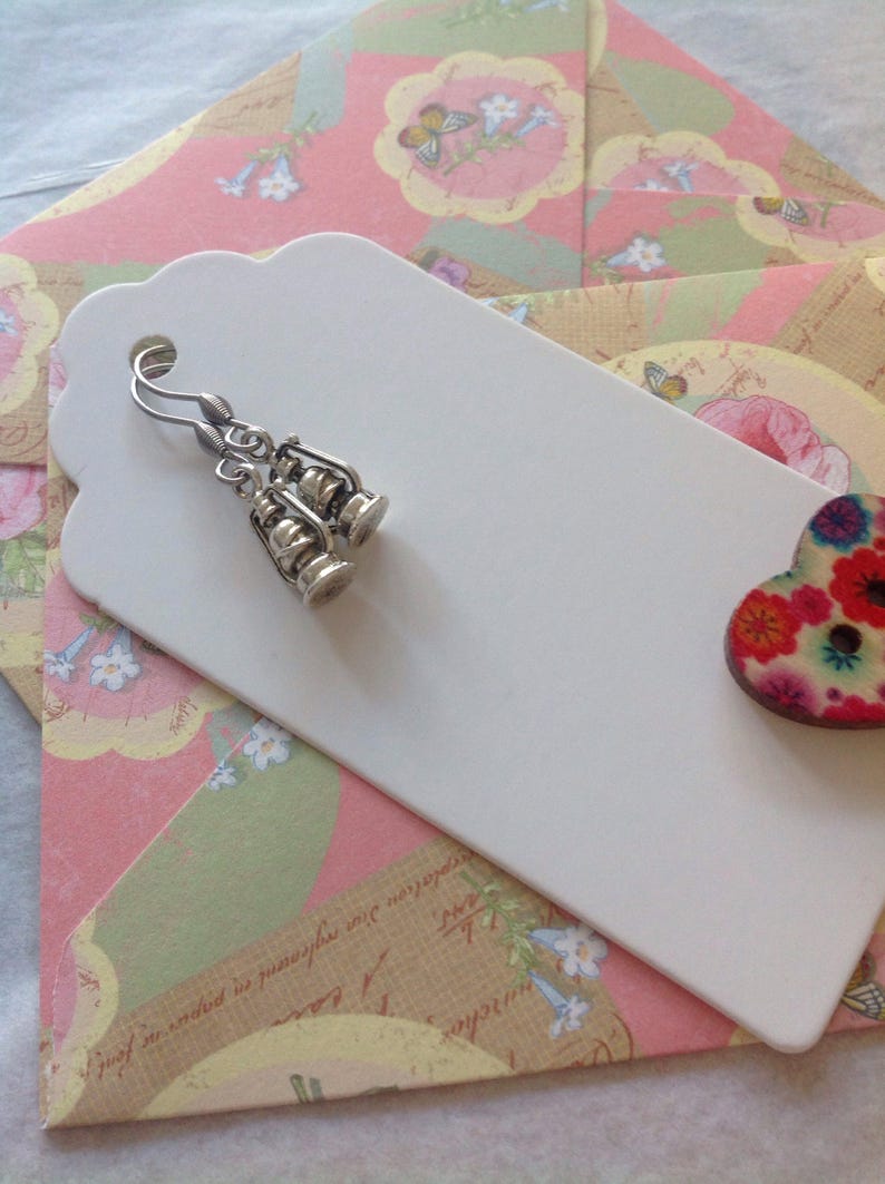 May include: A pair of silver earrings shaped like lanterns hanging from a white tag with a floral patterned envelope behind it.