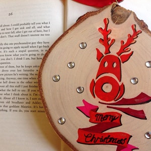 May include: A wooden Christmas ornament with a red reindeer silhouette and the words "Merry Christmas" in pink and red lettering. The ornament is decorated with silver glitter.