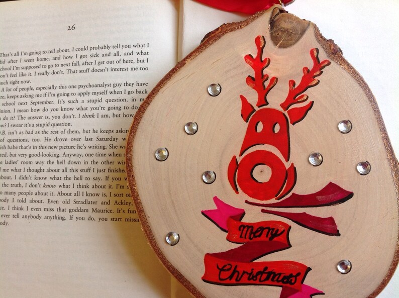 May include: A wooden Christmas ornament with a red reindeer silhouette and the words "Merry Christmas" in pink and red lettering. The ornament is decorated with silver glitter.