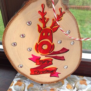 May include: A wooden Christmas ornament with a red reindeer painted on it. The ornament has a pink and red ribbon with the words "merry Christmas" written on it. The ornament is decorated with silver glitter.