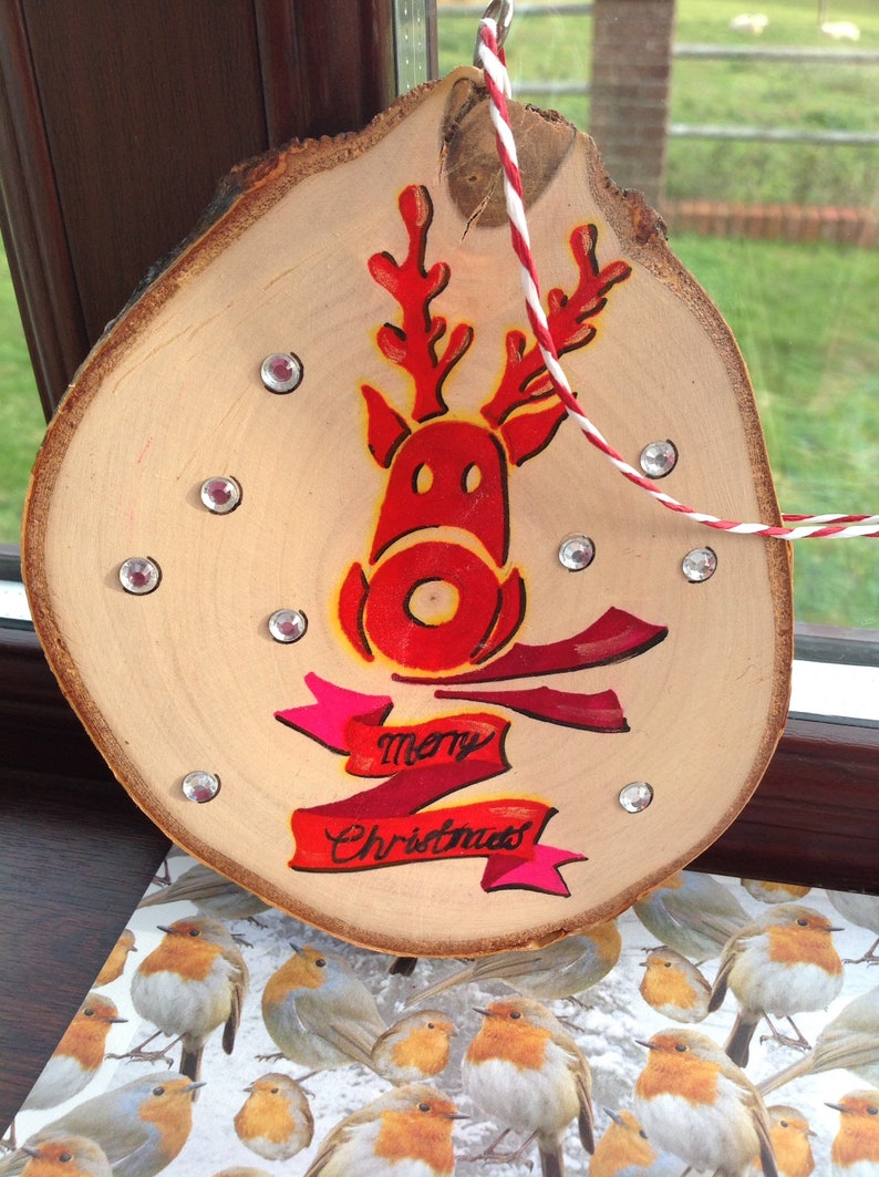 May include: A wooden Christmas ornament with a red reindeer painted on it. The ornament has a pink and red ribbon with the words "merry Christmas" written on it. The ornament is decorated with silver glitter.