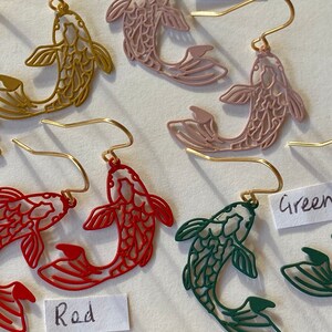 K&ouml;nnte beinhalten: Goldene fischf&ouml;rmige Ohrringe in Pink, Rot und Gr&uuml;n. Die Ohrringe sind ausgeschnitten und haben ein filigranes Design. Die Ohrringe sind mit den Farben "Pink", "Rot" und "Gr&uuml;n" beschriftet.