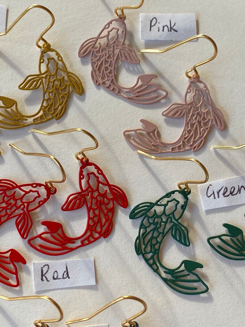 K&ouml;nnte beinhalten: Goldene fischf&ouml;rmige Ohrringe in Pink, Rot und Gr&uuml;n. Die Ohrringe sind ausgeschnitten und haben ein filigranes Design. Die Ohrringe sind mit den Farben "Pink", "Rot" und "Gr&uuml;n" beschriftet.
