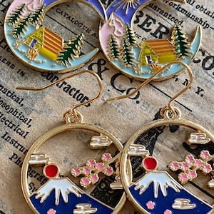 May include: A pair of gold-tone earrings featuring a crescent moon design with a cabin in the woods and a mountain scene with cherry blossoms. The earrings are made of metal and have a shiny finish.