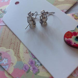 May include: A pair of silver earrings shaped like antique oil lamps. The earrings are on a white background.