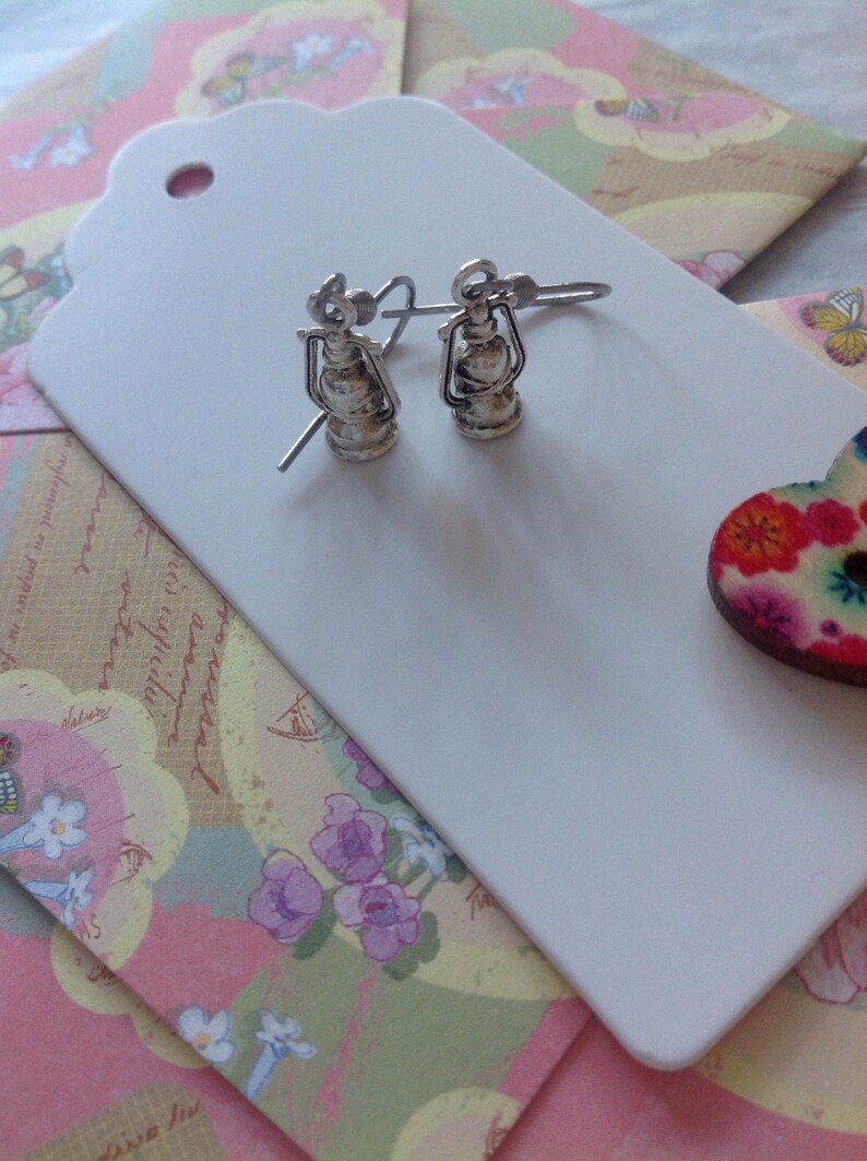 May include: A pair of silver earrings shaped like antique oil lamps. The earrings are on a white background.