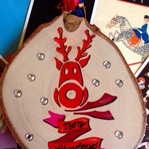 May include: A wooden Christmas ornament with a red reindeer and the words "merry Christmas" painted on it. The ornament is decorated with silver glitter.