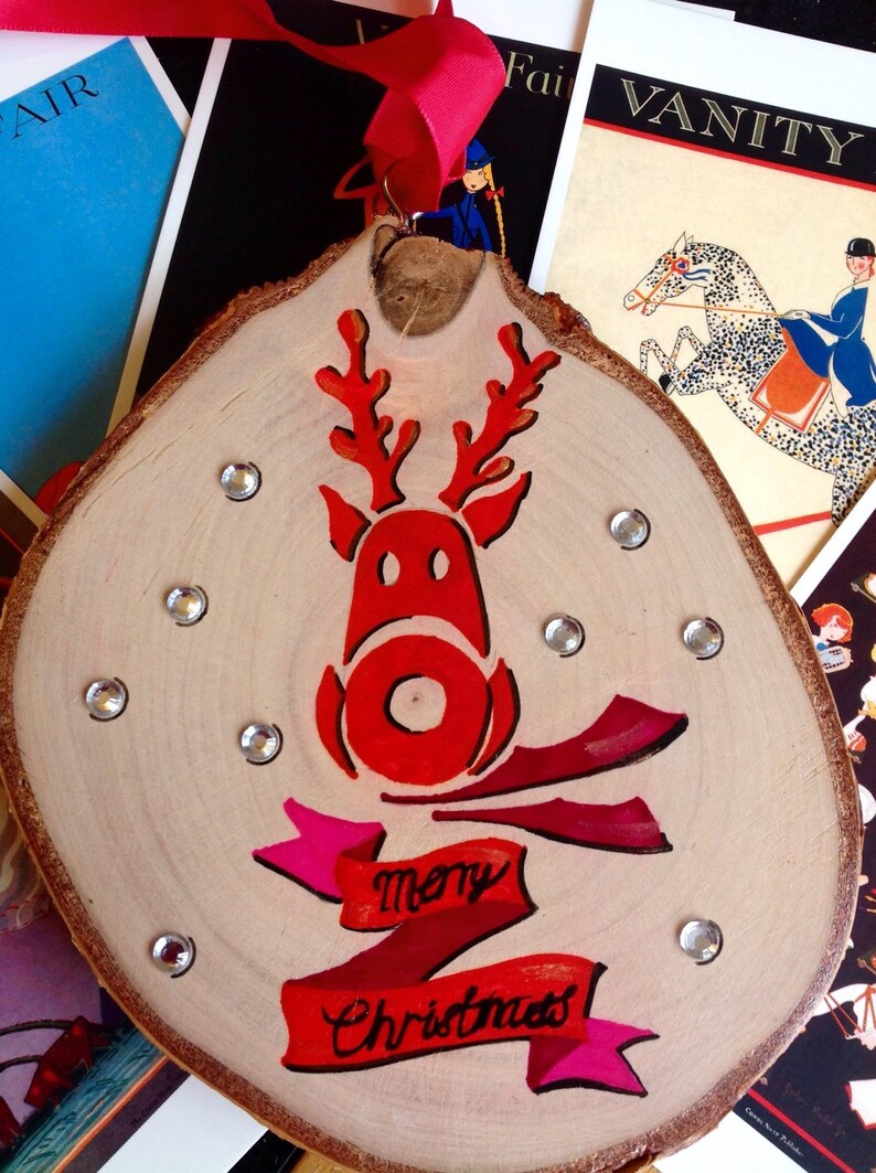 May include: A wooden Christmas ornament with a red reindeer and the words "merry Christmas" painted on it. The ornament is decorated with silver glitter.