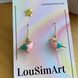 May include: A pair of gold earrings featuring a pink and turquoise bird design. The earrings are displayed on a white card with the text "LouSimArt" and "Lousimart.Etsy.com".