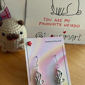 May include: Silver-toned earrings featuring a cup design filled with scissors and paintbrushes. The earrings hang from a silver hook. A card with the text "You are my favourite weirdo" and a crocheted pug are in the background.