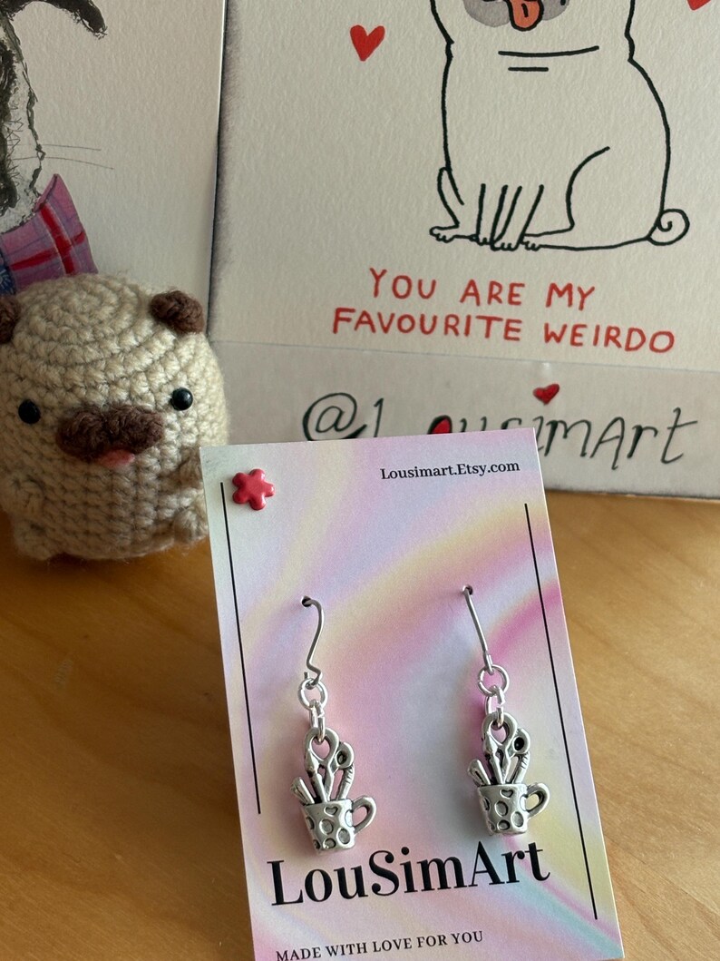 May include: Silver-toned earrings featuring a cup design filled with scissors and paintbrushes. The earrings hang from a silver hook. A card with the text "You are my favourite weirdo" and a crocheted pug are in the background.