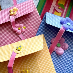 Op de afbeelding: Vier kleurrijke enveloppen met ruitjespatronen en decoratieve versieringen, waaronder bloemen en harten. De enveloppen zijn vastgebonden met een roze lint.