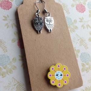 Pode incluir: Um par de brincos de prata em forma de coruja com um bot&atilde;o de flor amarelo com detalhes em rosa e azul.