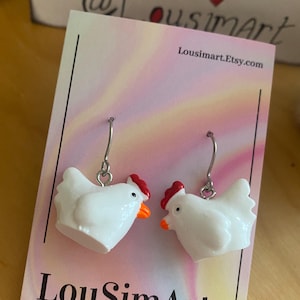 Puede incluir: Pendientes de pollo de cerámica blanca con detalles rojos y naranjas. Los pendientes están colgados de una tarjeta blanca con el texto "LouSimArt" y "Made with love for you".