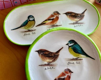May include: Two white ceramic trays with green rims, hand-painted with colorful bird illustrations. The oval tray features three birds, while the round tray displays three different birds. Text includes "Blue tit", "Wren", and "Chaffinch".