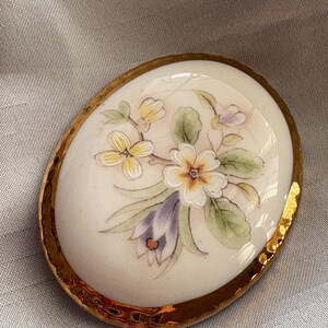 Może przedstawiać: Owalna broszka porcelanowa ze złotą, teksturowaną obw&oacute;dką. Broszka przedstawia ręcznie malowany kwiatowy wz&oacute;r z białymi, ż&oacute;łtymi i fioletowymi kwiatami i zielonymi liśćmi. Tło jest kremowo-białe.