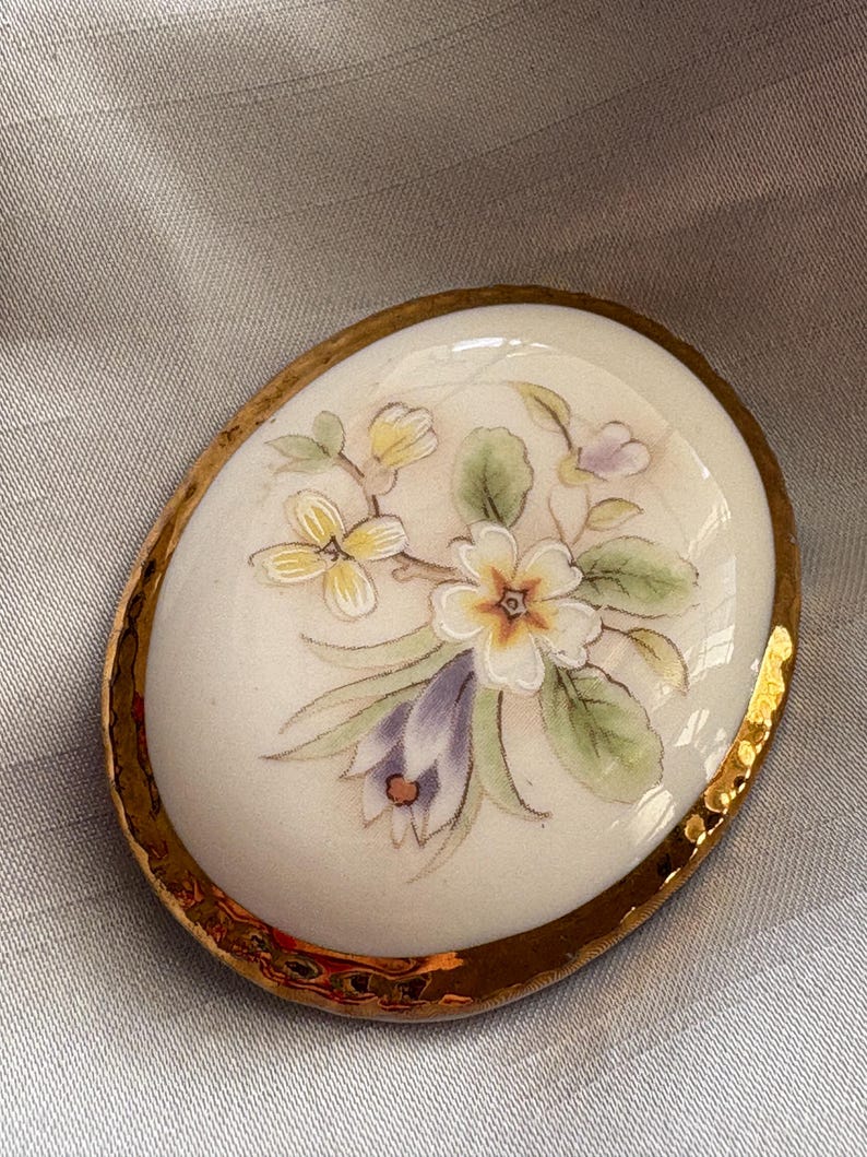 Może przedstawiać: Owalna broszka porcelanowa ze złotą, teksturowaną obw&oacute;dką. Broszka przedstawia ręcznie malowany kwiatowy wz&oacute;r z białymi, ż&oacute;łtymi i fioletowymi kwiatami i zielonymi liśćmi. Tło jest kremowo-białe.