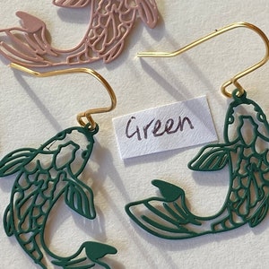 K&ouml;nnte beinhalten: Ein Paar goldene Ohrringe mit gr&uuml;nen und rosa Fisch-Anh&auml;ngern. Die Fisch-Anh&auml;nger sind ausgeschnitten und haben ein detailliertes Design. Das Wort "Gr&uuml;n" ist auf einem wei&szlig;en Papier geschrieben.