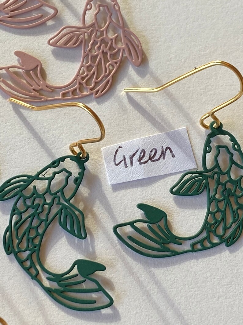K&ouml;nnte beinhalten: Ein Paar goldene Ohrringe mit gr&uuml;nen und rosa Fisch-Anh&auml;ngern. Die Fisch-Anh&auml;nger sind ausgeschnitten und haben ein detailliertes Design. Das Wort "Gr&uuml;n" ist auf einem wei&szlig;en Papier geschrieben.