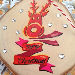 May include: A wooden Christmas ornament with a red reindeer painted on it. The reindeer has a red nose and antlers. The ornament also has a red and pink ribbon with the words "Merry Christmas" written on it.