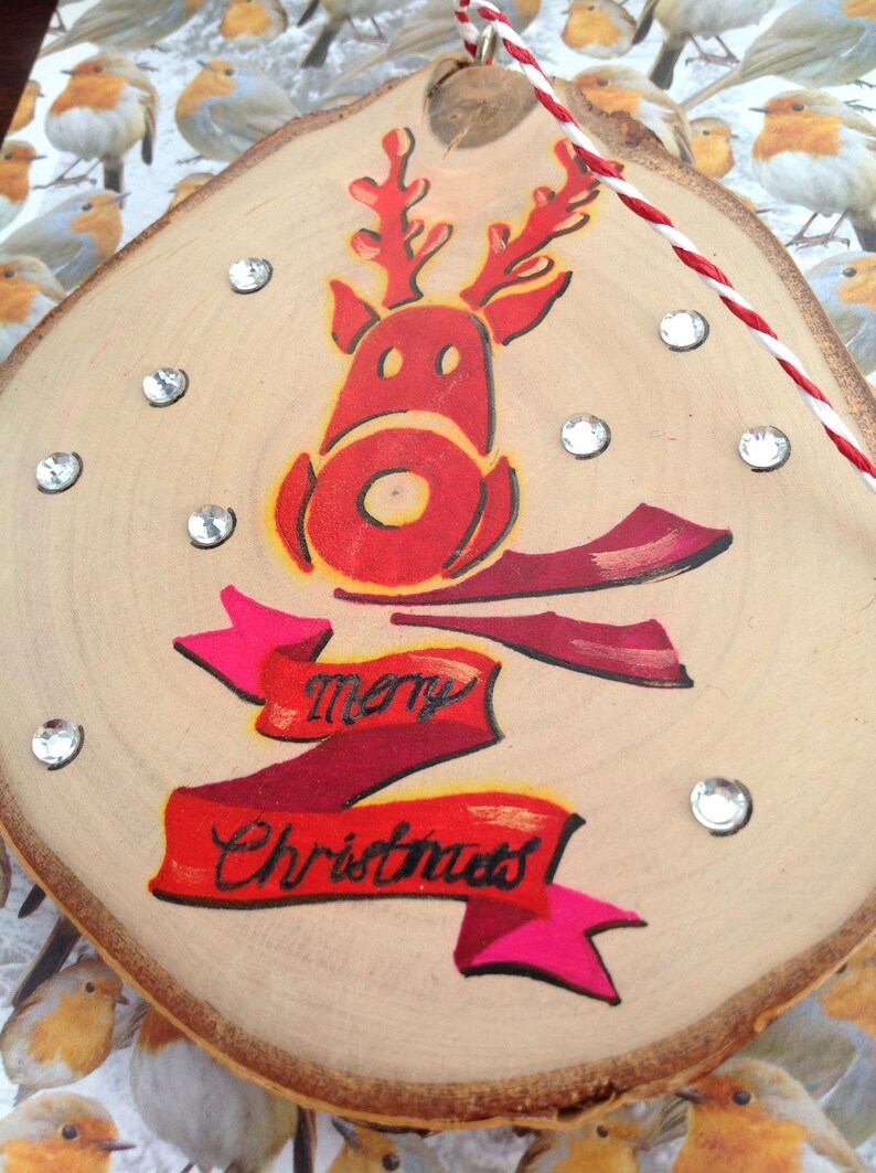 May include: A wooden Christmas ornament with a red reindeer painted on it. The reindeer has a red nose and antlers. The ornament also has a red and pink ribbon with the words "Merry Christmas" written on it.