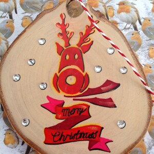 May include: A wooden Christmas ornament with a red reindeer painted on it. The reindeer has a red nose and antlers. The ornament also has a red and pink ribbon with the words "Merry Christmas" written on it.