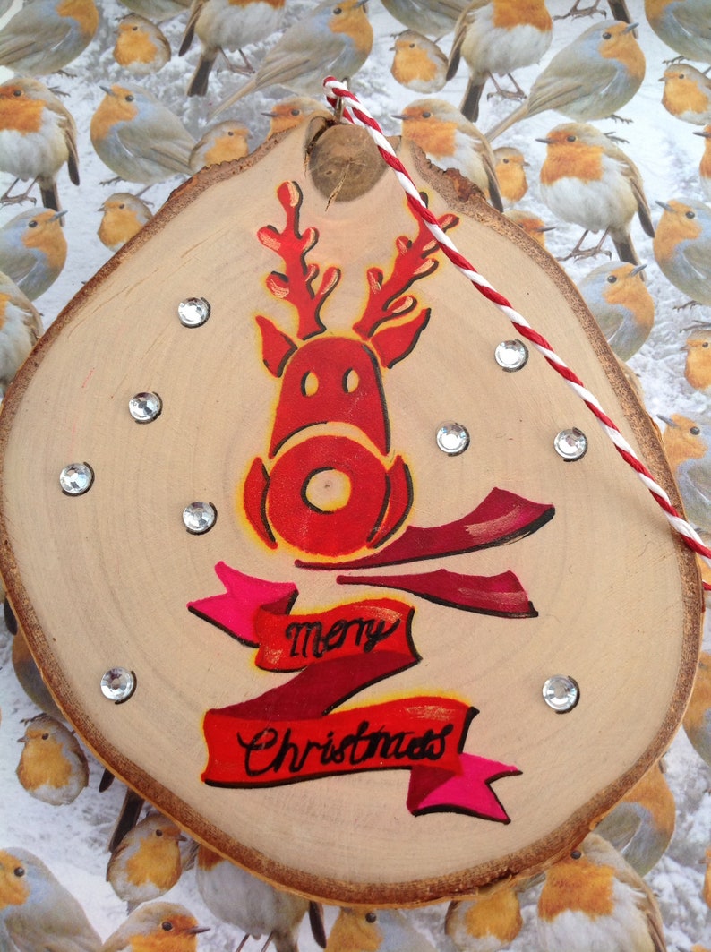 May include: A wooden Christmas ornament with a red reindeer painted on it. The reindeer has a red nose and antlers. The ornament also has a red and pink ribbon with the words "Merry Christmas" written on it.