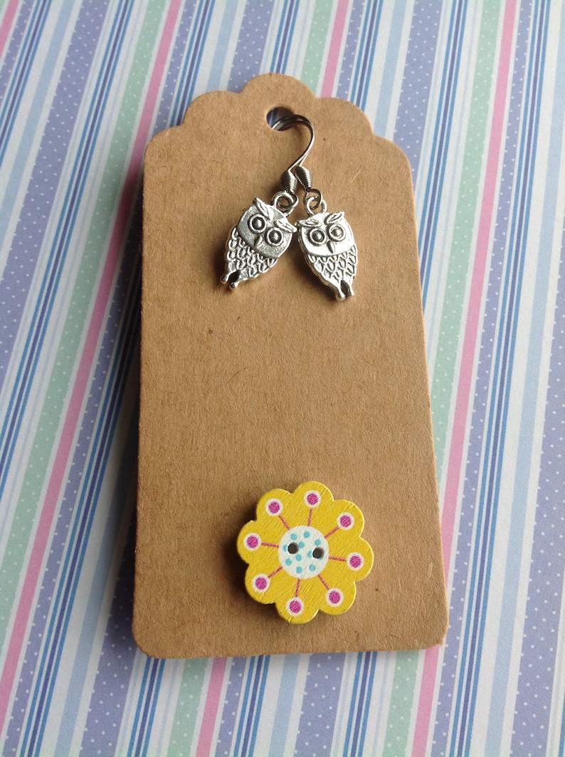 Pode incluir: Uma etiqueta de papel castanho com brincos de coruja prateados e um bot&atilde;o de flor amarelo com detalhes rosa e azul.