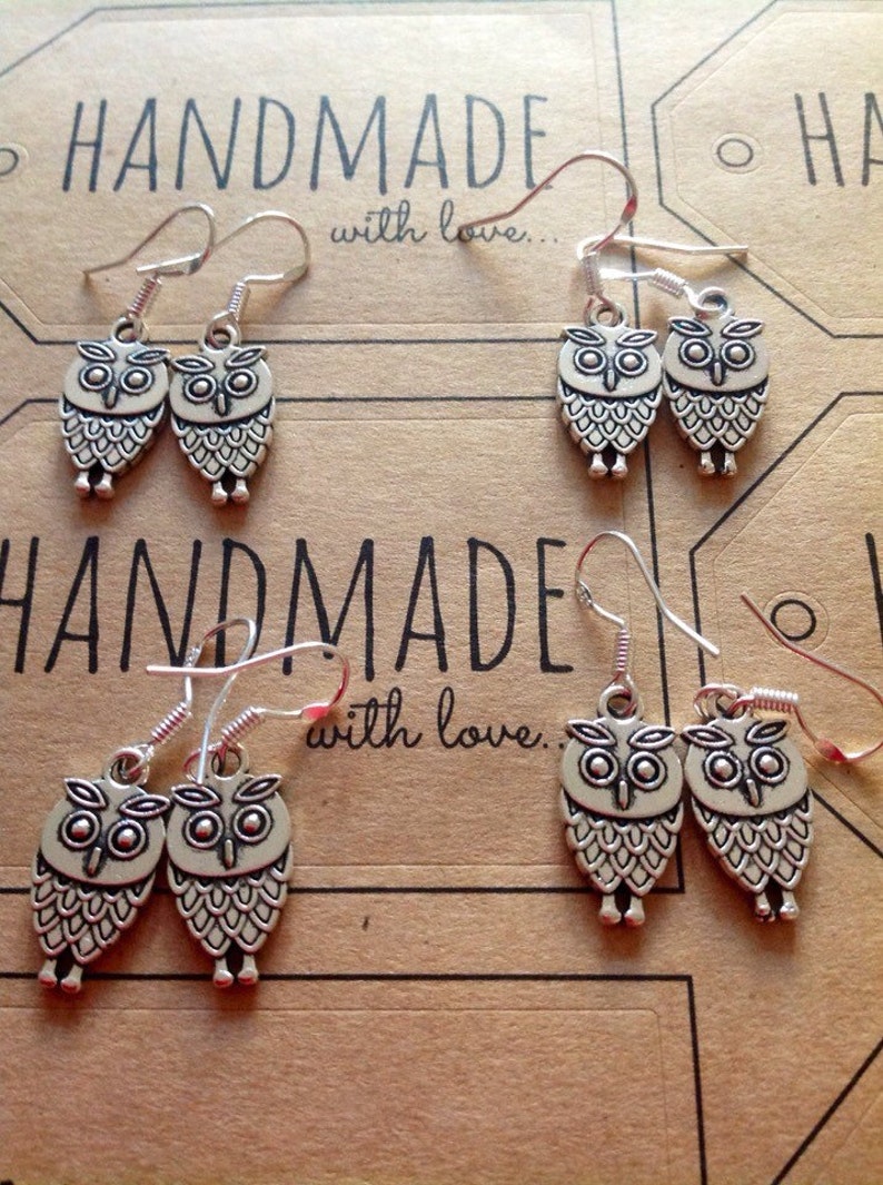 Pode incluir: Tr&ecirc;s pares de brincos de prata com formato de coruja com fecho de gancho. Os brincos s&atilde;o feitos &agrave; m&atilde;o e apresentam o texto "Handmade with love." no fundo.