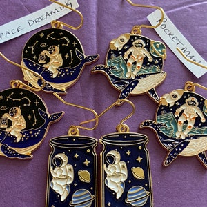 Puede incluir: Un conjunto de cuatro pendientes dorados con diseños de astronautas y temática espacial. Los pendientes tienen forma de ballena, frasco y cohete. Los pendientes de ballena presentan una ballena azul y negra con estrellas y constelaciones. Los pendientes de frasco presentan un frasco azul y negro con un astronauta flotando dentro. Los pendientes de cohete presentan un cohete azul y negro con un astronauta flotando fuera. Los pendientes están etiquetados como "Space Dreaming", "Rocket Man" y "Space Jar".