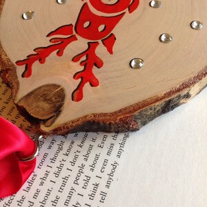 May include: A wooden ornament with a red reindeer design and rhinestones. The ornament is attached to a red ribbon with a metal ring.