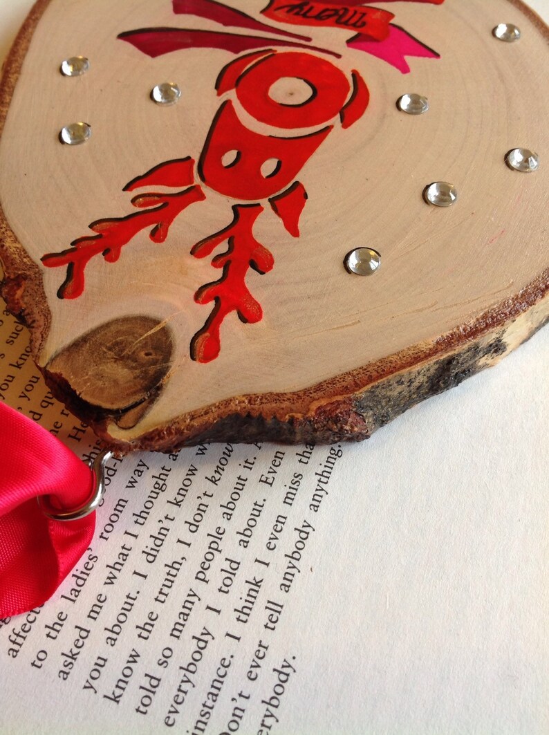 May include: A wooden ornament with a red reindeer design and rhinestones. The ornament is attached to a red ribbon with a metal ring.