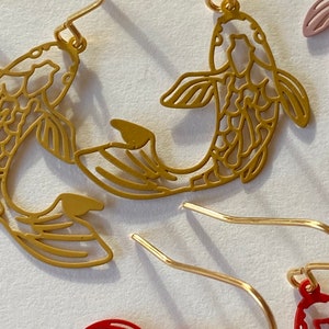 K&ouml;nnte beinhalten: Ein Paar goldfarbene Ohrringe mit einem filigranen, ausgeschnittenen Design aus zwei Koi-Fischen. Die Fische sind einander zugewandt und haben auf ihren ganzen K&ouml;rpern komplizierte Details.