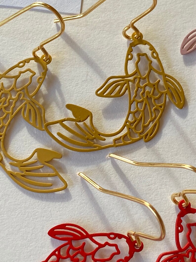 K&ouml;nnte beinhalten: Ein Paar goldfarbene Ohrringe mit einem filigranen, ausgeschnittenen Design aus zwei Koi-Fischen. Die Fische sind einander zugewandt und haben auf ihren ganzen K&ouml;rpern komplizierte Details.