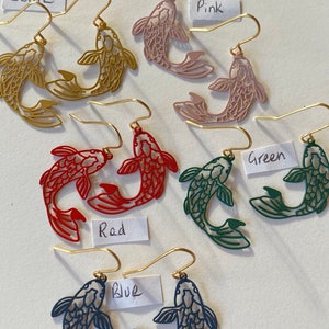 K&ouml;nnte beinhalten: Ein Set mit sechs Paaren goldfarbener Fische-Ohrringe in verschiedenen Farben, darunter Rot, Blau, Gr&uuml;n, Pink und Ocker. Jedes Paar zeigt zwei Fische, die sich gegen&uuml;berstehen und ein symmetrisches Design bilden.