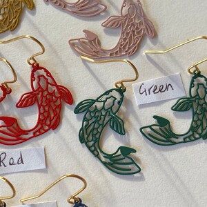 K&ouml;nnte beinhalten: Ein Set mit vier Paaren goldener Fische als Ohrringe. Die Fische sind aus Metall ausgeschnitten und in Rot, Rosa, Gr&uuml;n und Blau erh&auml;ltlich.
