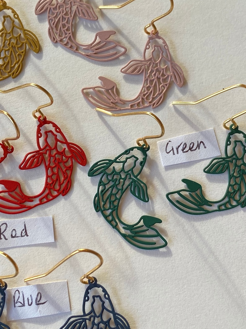 K&ouml;nnte beinhalten: Ein Set mit vier Paaren goldener Fische als Ohrringe. Die Fische sind aus Metall ausgeschnitten und in Rot, Rosa, Gr&uuml;n und Blau erh&auml;ltlich.